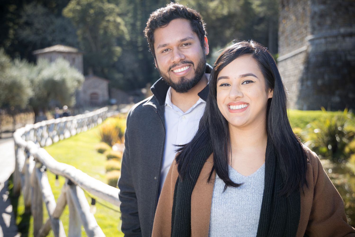 A couple poses together on a trail with a rustic bridge and scenic landscape behind them.