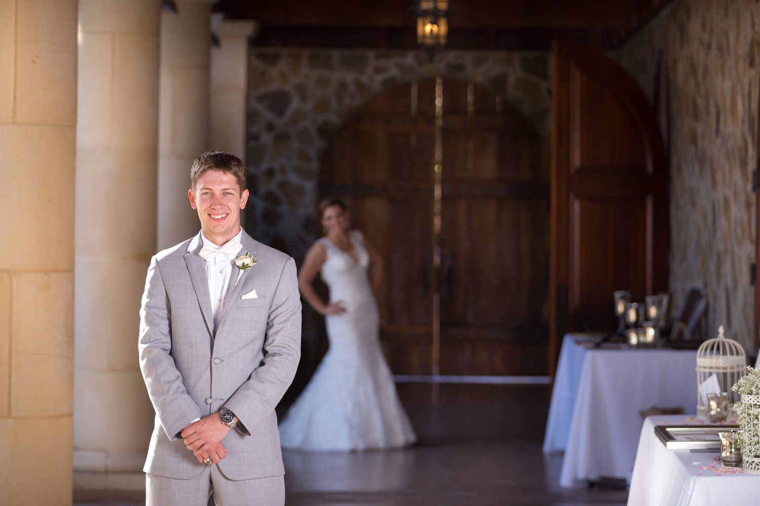 Groom in light gray suit standing in stone archway with reception tables visible.