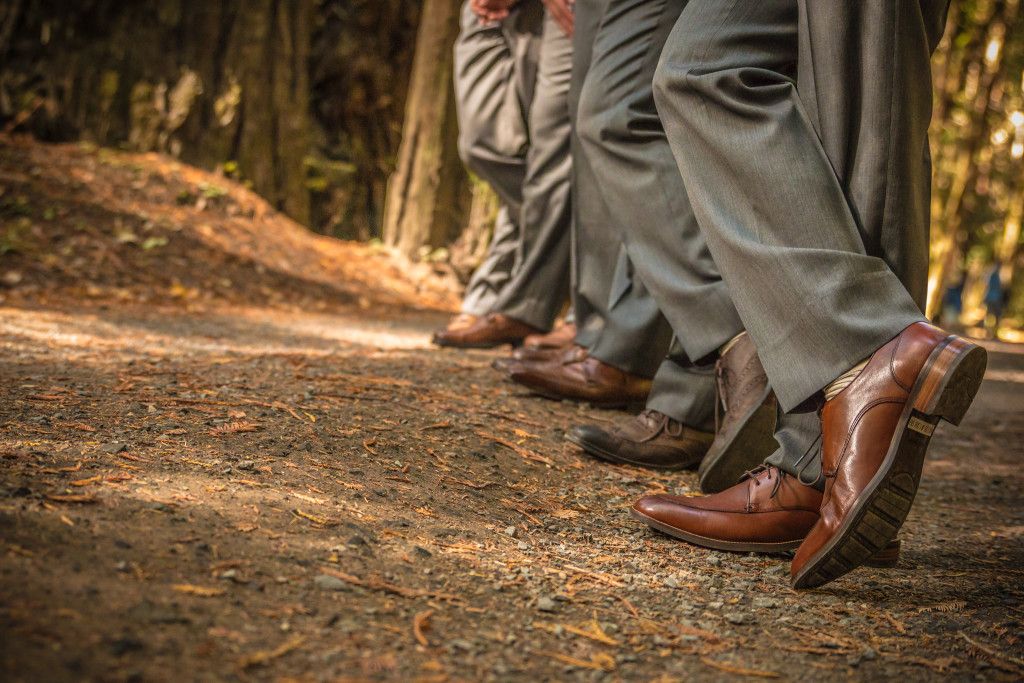 Line of brown leather dress shoes and grey suit pants on a forest trail.