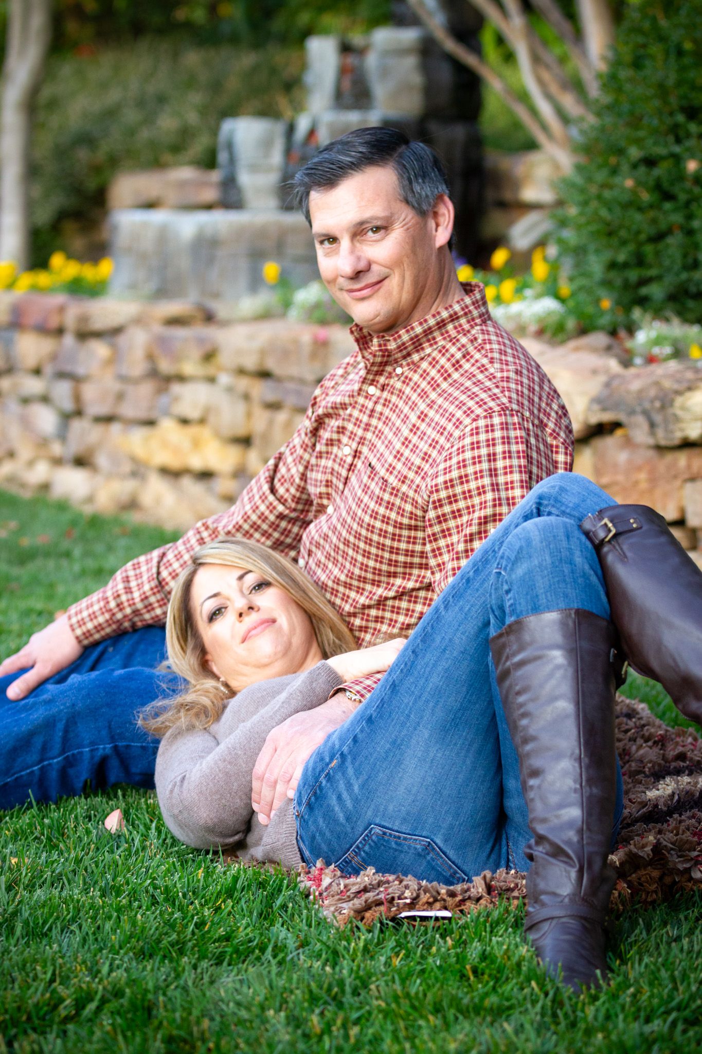 Couple relaxing together on grass by stone wall in garden during outdoor photoshoot.