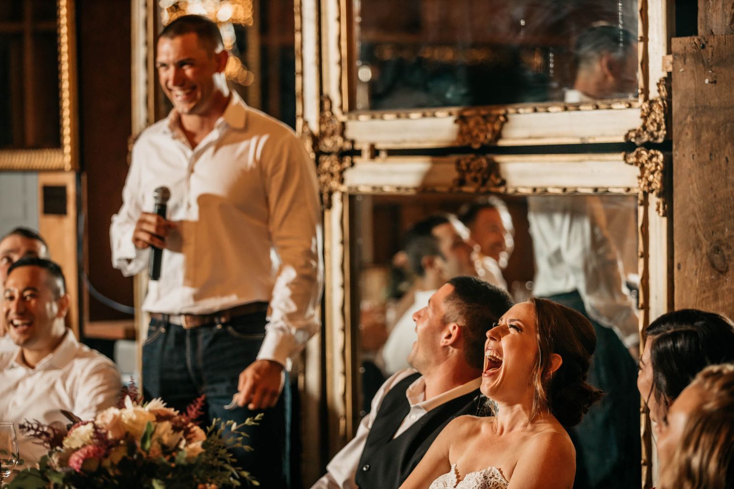 Wedding guests laughing during speeches at a reception with ornate mirrored walls.