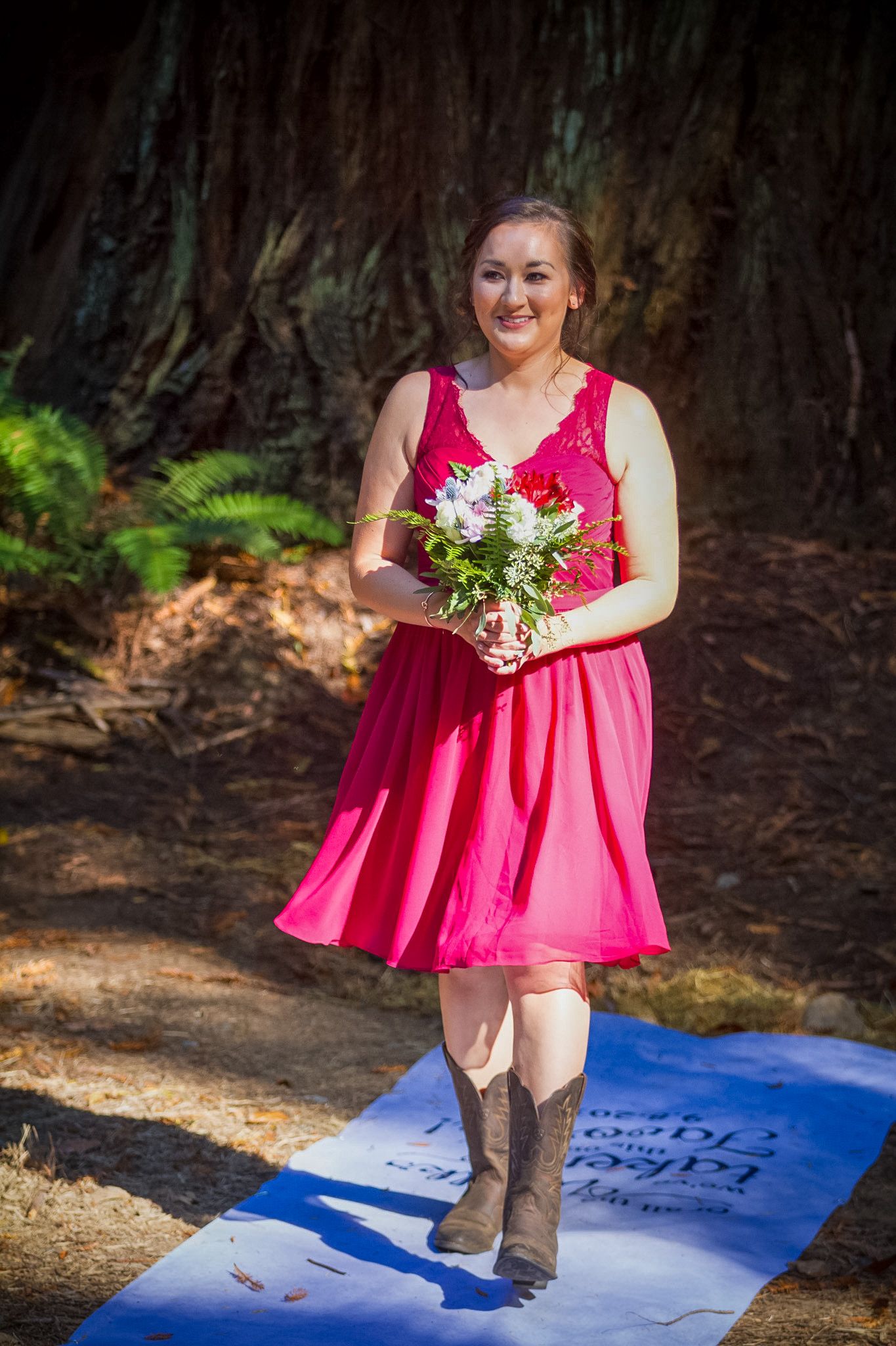Bridesmaid in red dress and cowboy boots walking on aisle runner in redwood forest.