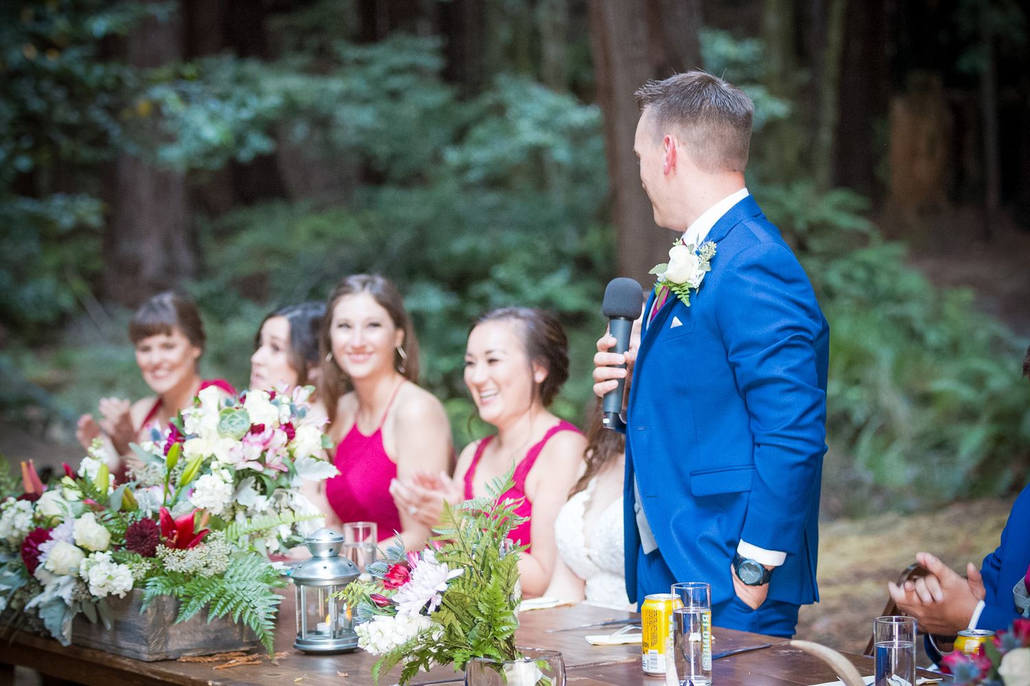 Wedding reception table in forest setting with guests and floral centerpieces.