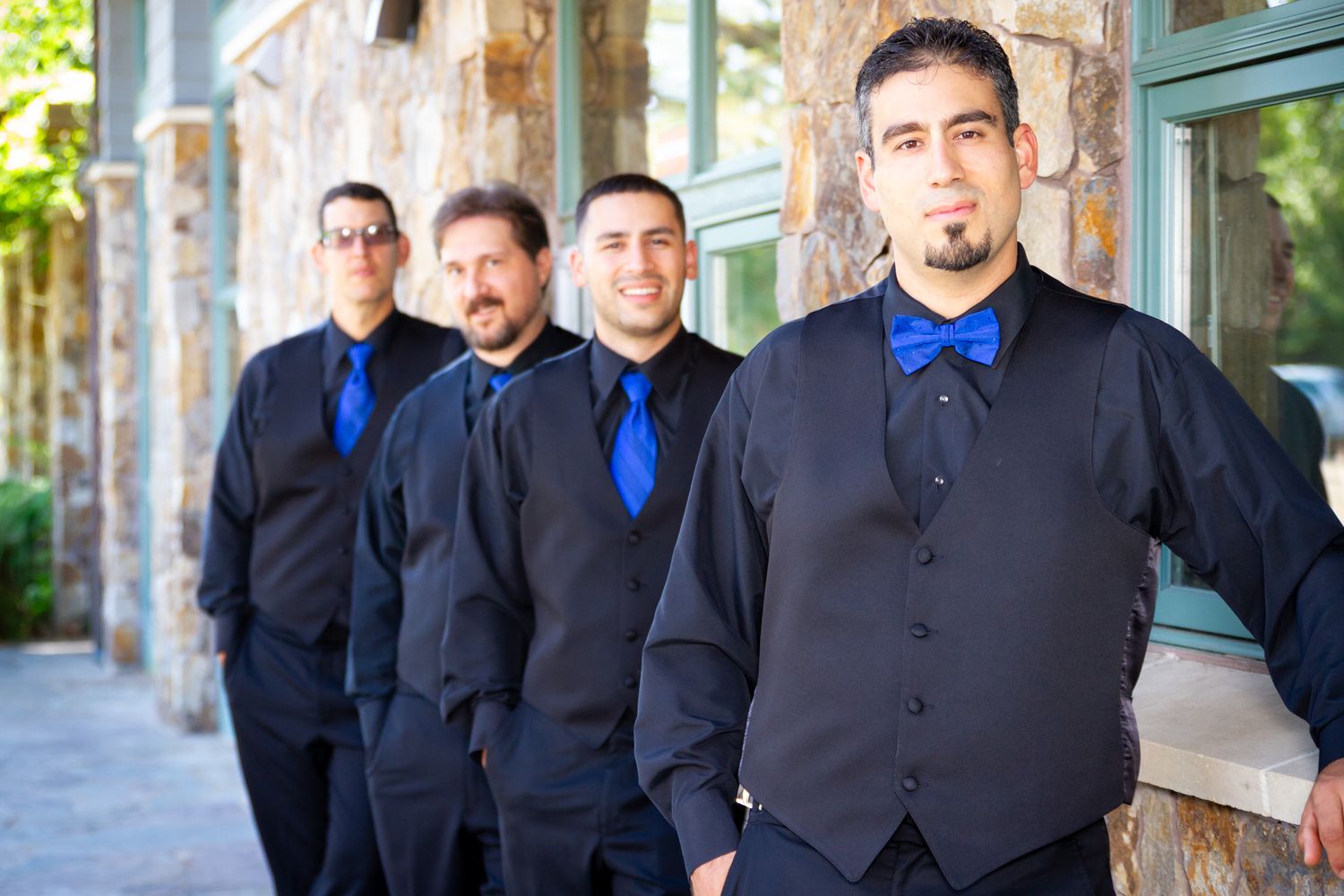 Groomsmen in matching black suits with blue ties stand along stone wall.