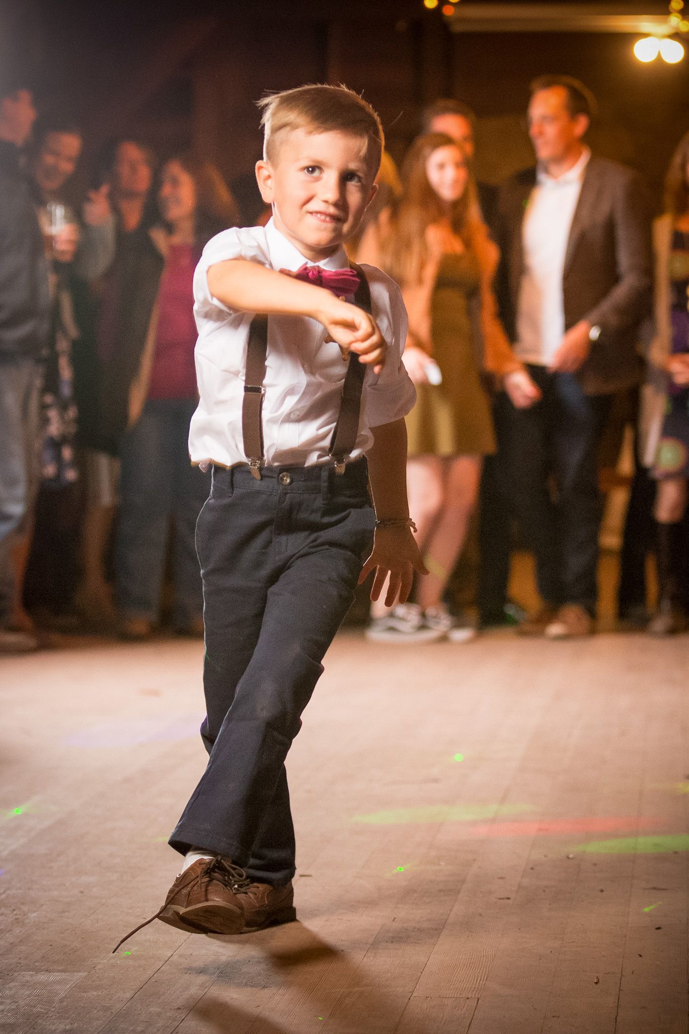 A young boy in suspenders and bow tie dances at a celebration with party guests watching in the background.