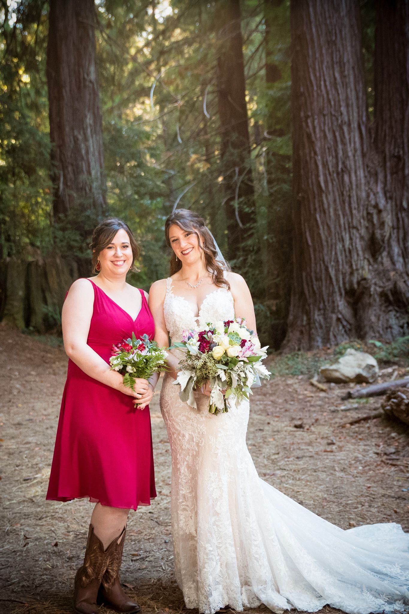 Bride and bridesmaid with floral bouquets stand together on forest path.