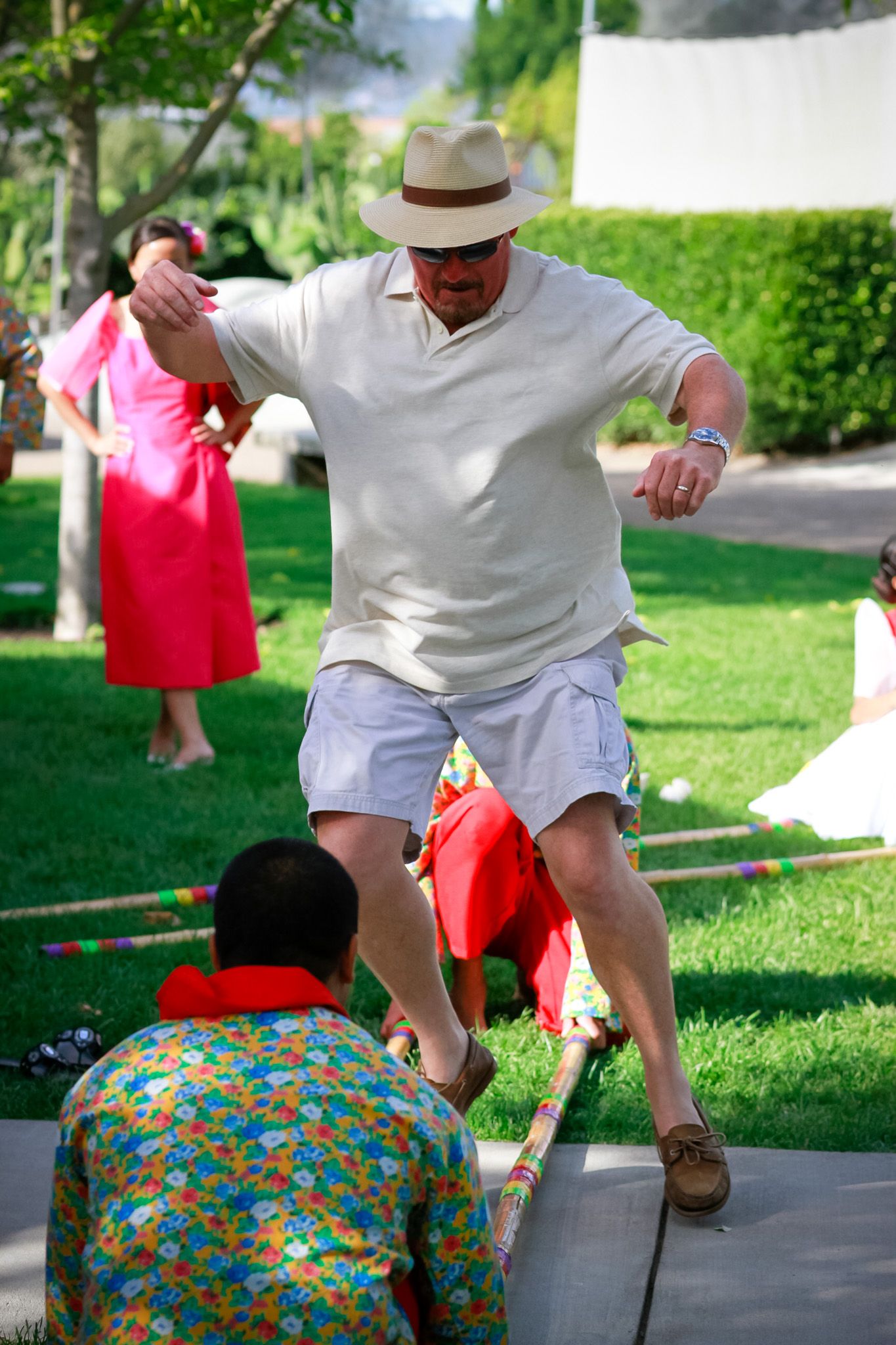 Performer in white clothing and hat jumping over bamboo poles during outdoor event.