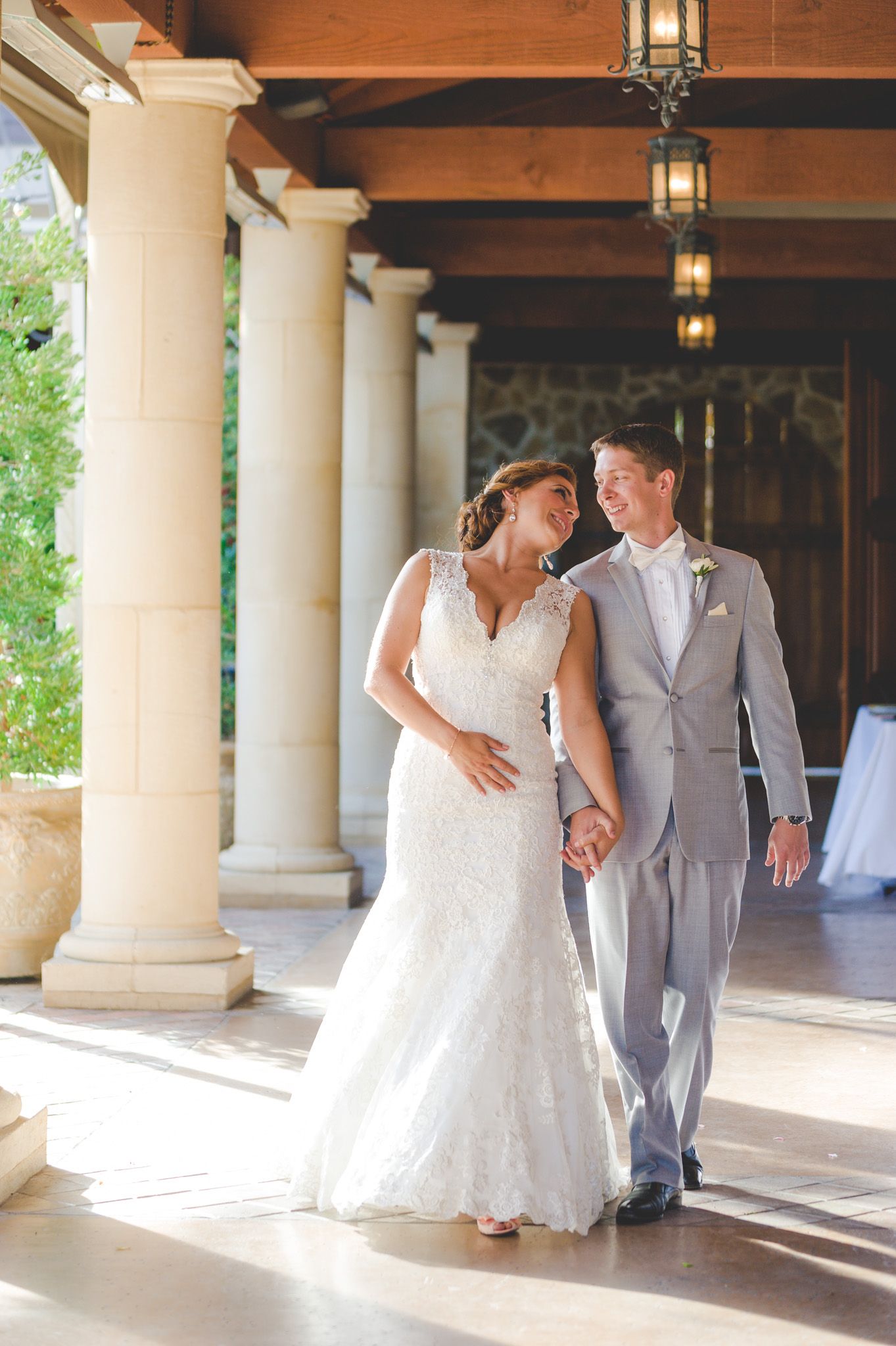 A couple in wedding attire share a romantic moment while walking under a covered porch with elegant stone columns.