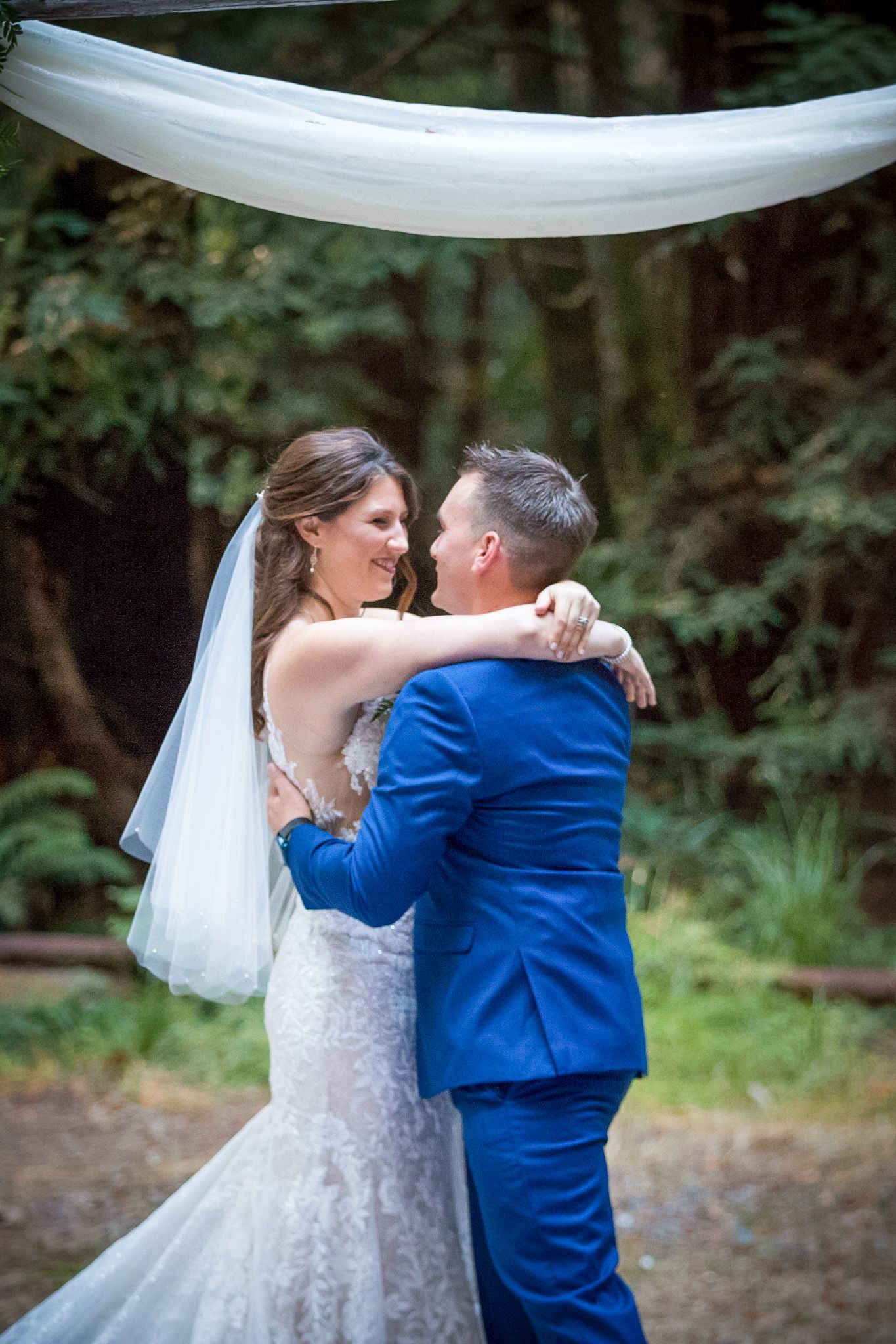 Newlyweds share their first dance under white fabric draping in a forest setting.