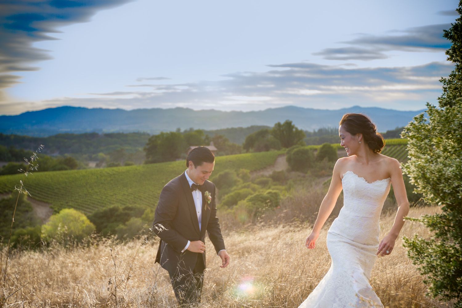 Couple walking through golden grass field with mountains in background.