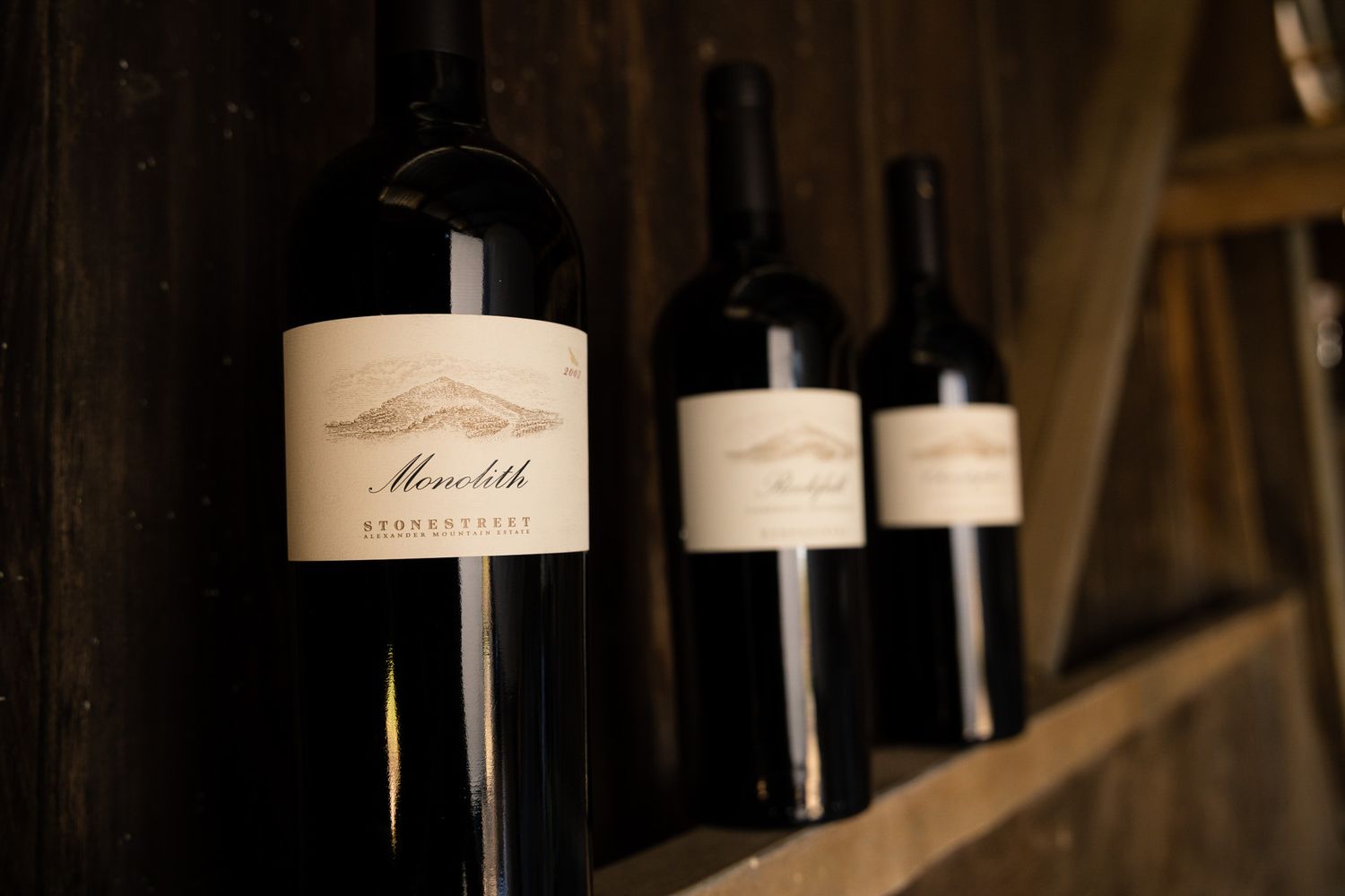Three wine bottles with elegant white labels displayed on wooden shelving in a cellar.