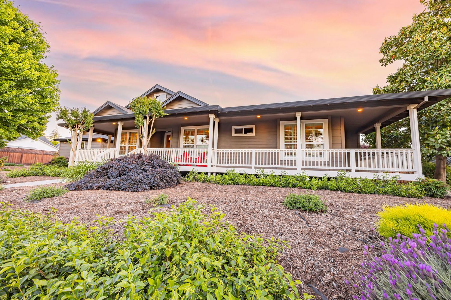 Beautiful craftsman style home with wraparound porch and landscaped front yard with purple flowers at dusk.