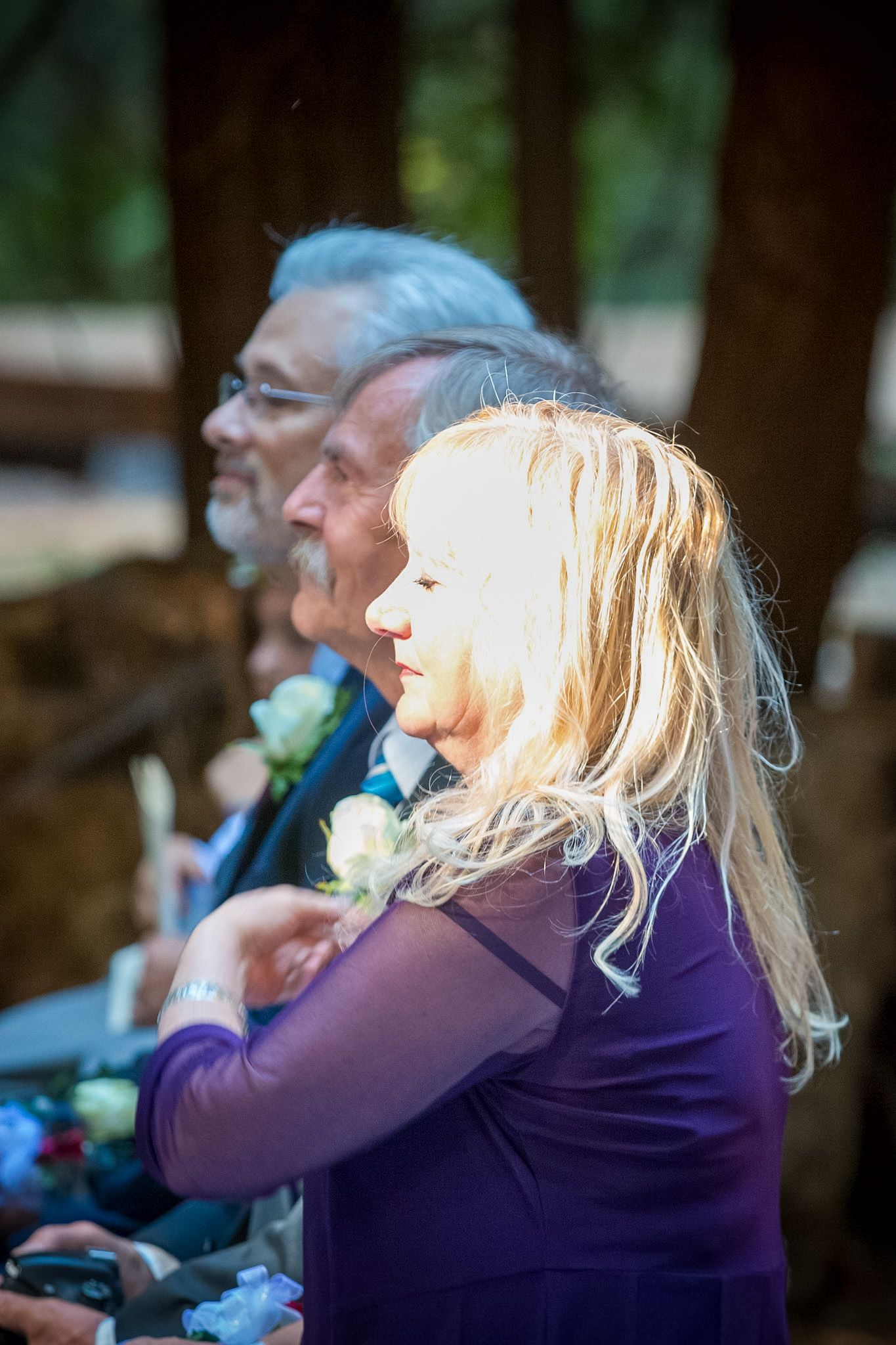 Three guests in formal attire sitting together at an outdoor wedding ceremony with white boutonnieres.