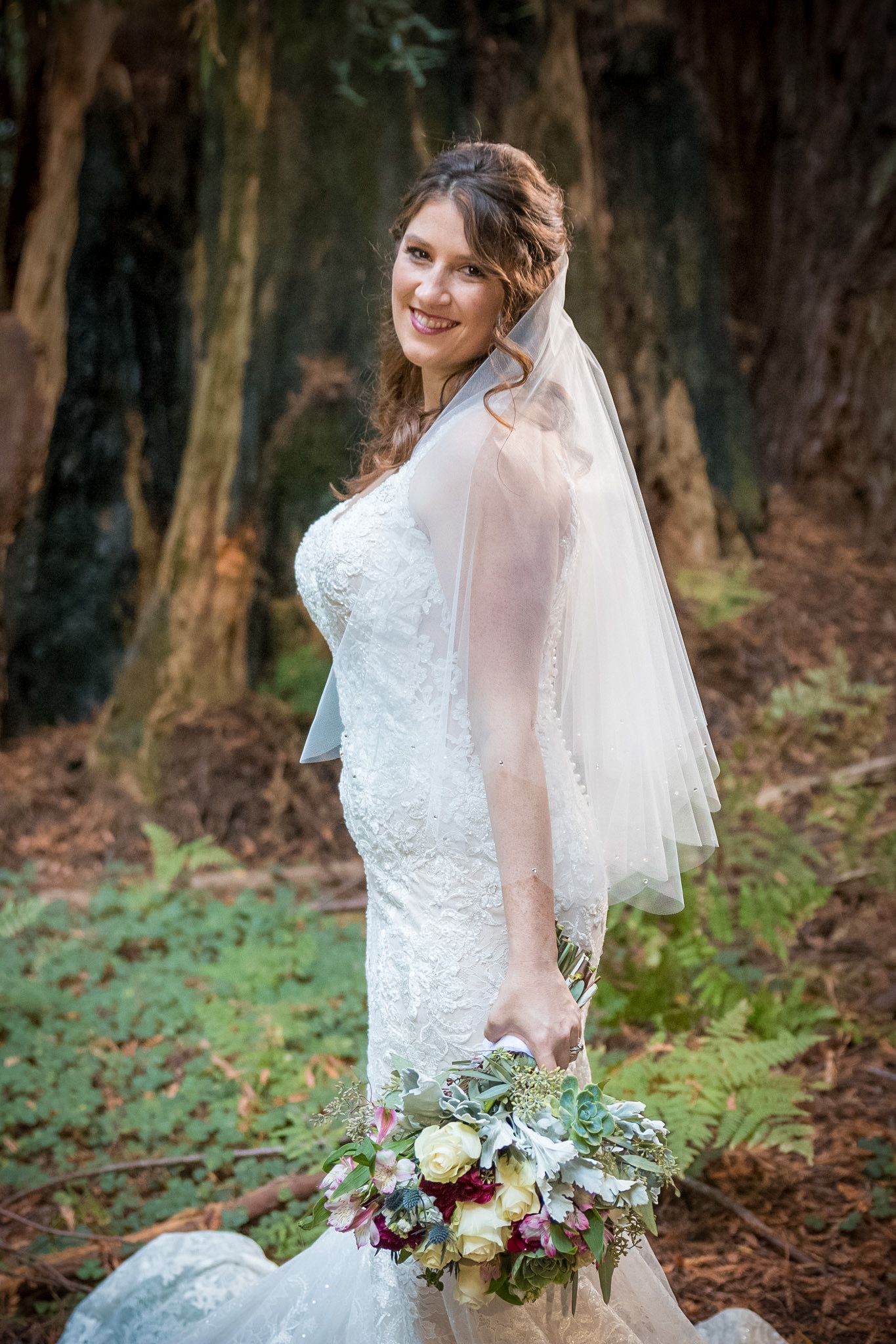 Bride wearing a fitted lace gown and veil poses with her bouquet in a wooded outdoor setting.