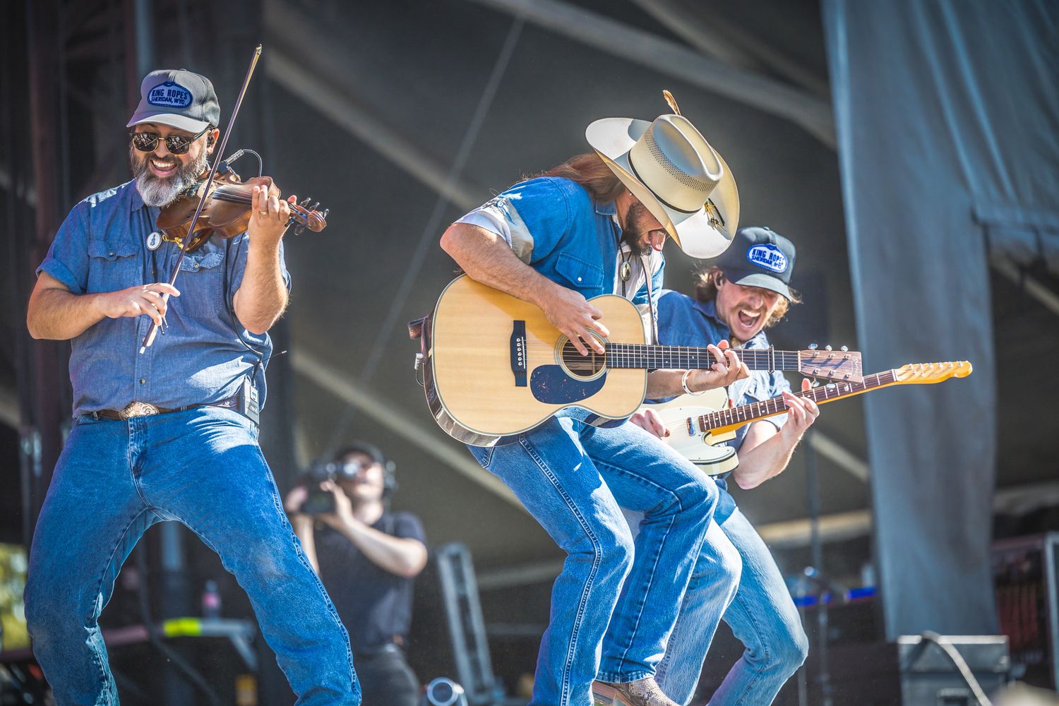 Musicians in blue outfits perform energetically on stage with violin and acoustic guitar during outdoor concert.