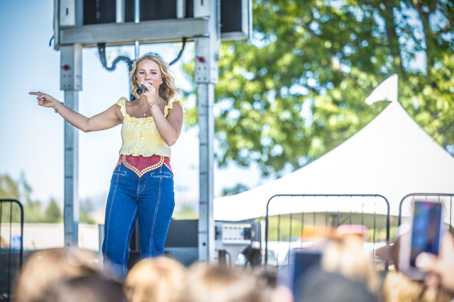 A performer sings on an outdoor stage during a sunny summer day with trees in the background.