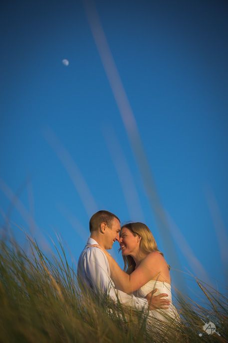Romantic couple embracing in tall grass against blue sky with moon visible at dusk.