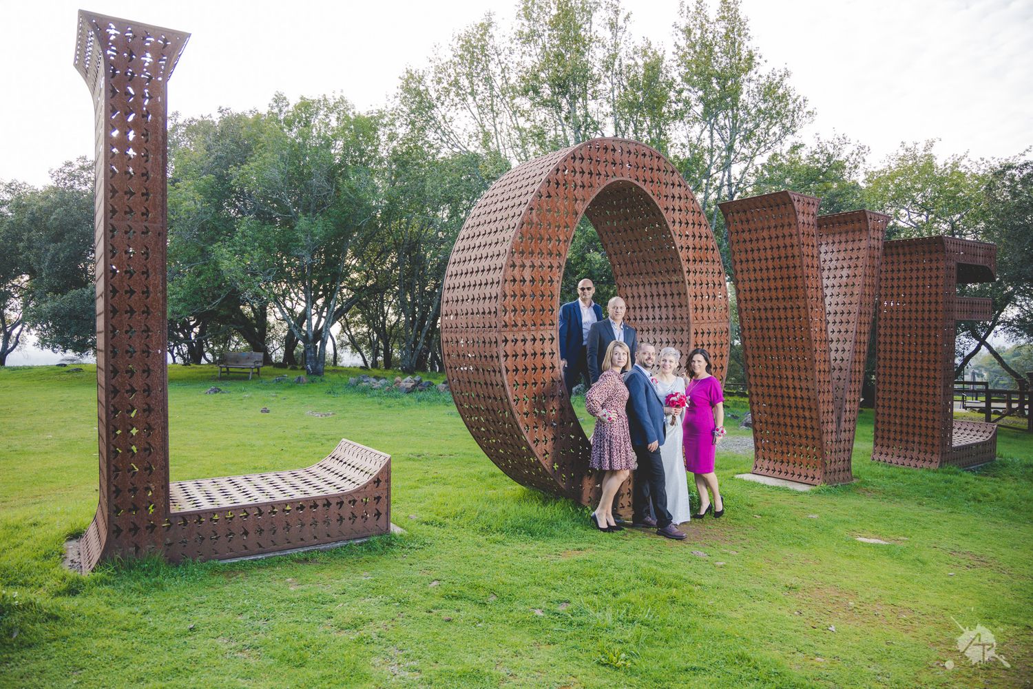 Outdoor art installation with curved brick structures spelling 'LOVE' on a grassy lawn with trees in the background.