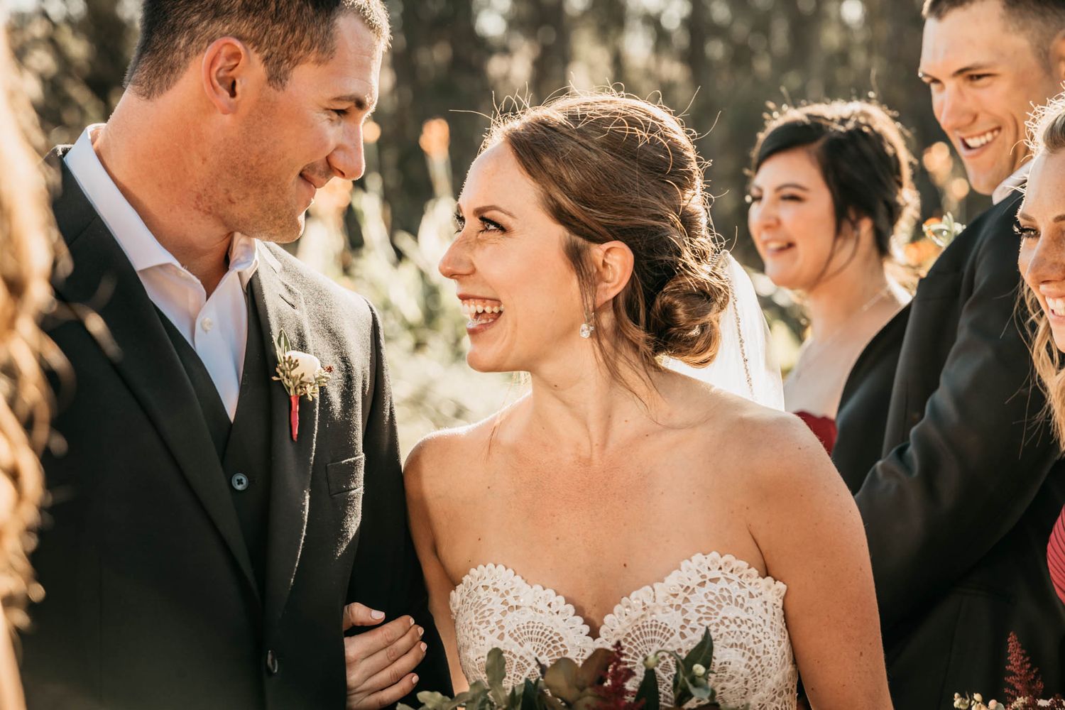 A candid wedding moment captured in warm sunlight with a laughing couple surrounded by their wedding party.