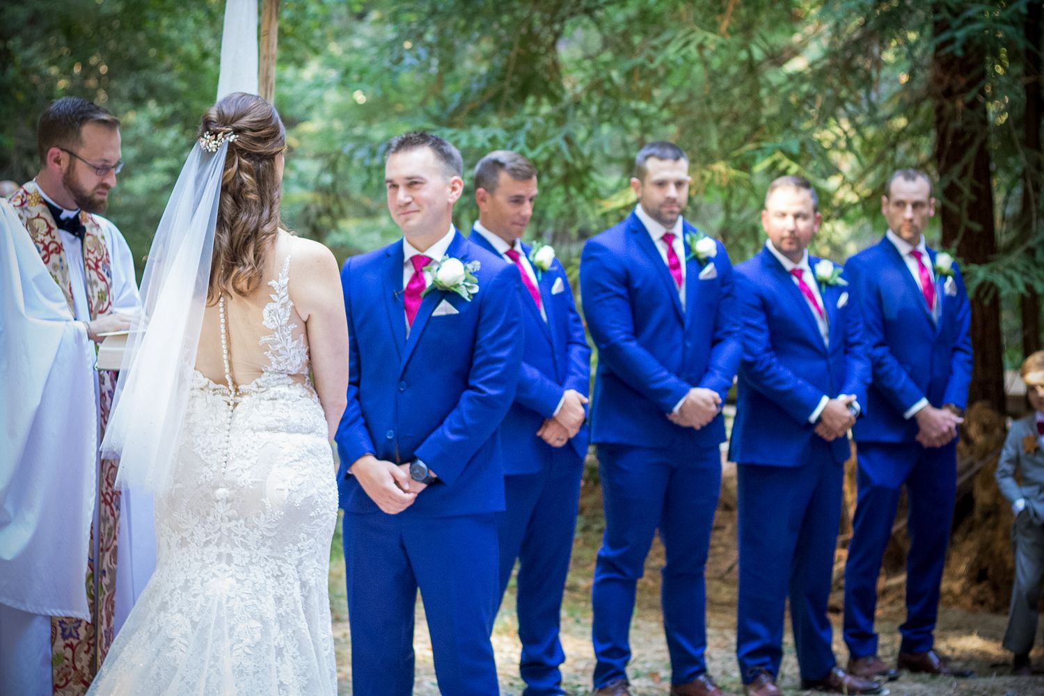 A wedding party lined up during ceremony with groomsmen wearing bright blue suits in a forest setting.