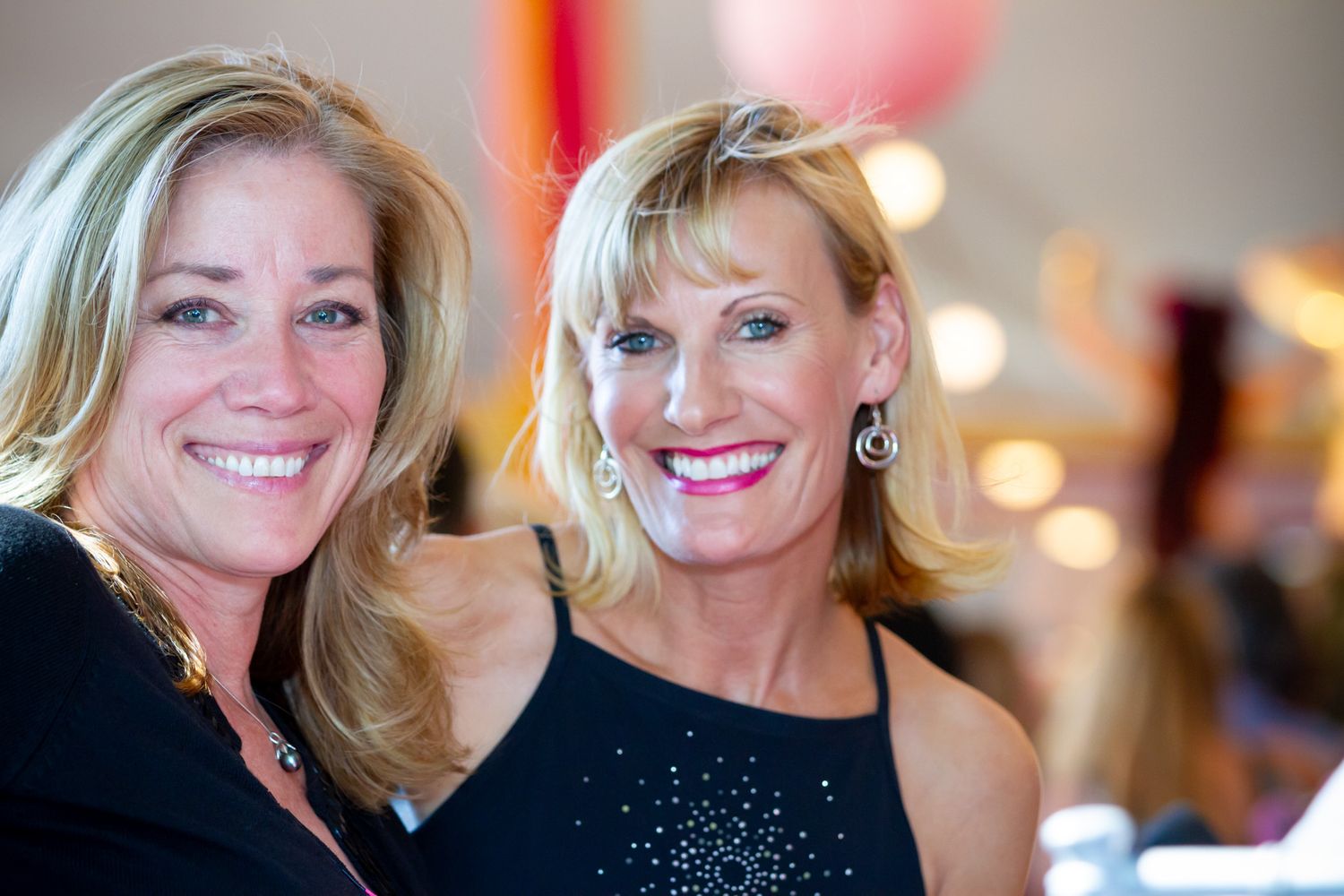 Two women smile warmly at a social gathering wearing black formal attire.