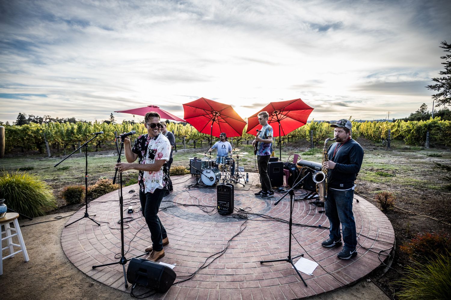 Band performs on circular stage under red umbrellas at outdoor vineyard venue during cloudy afternoon.