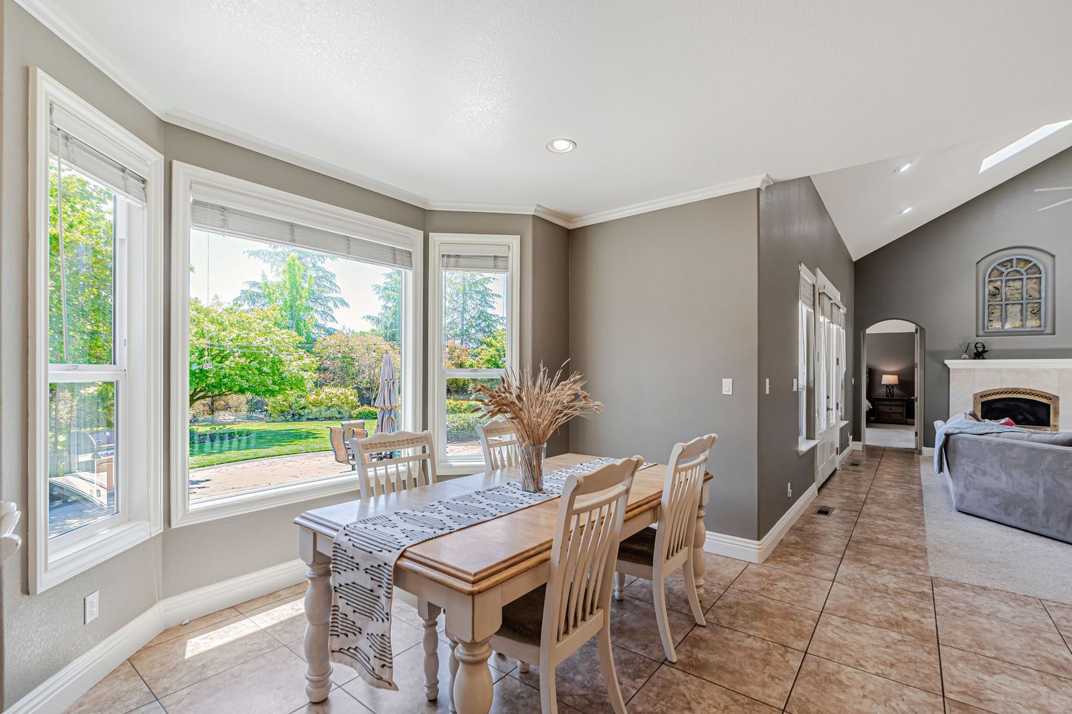 Bright dining area with bay windows, rustic white table and chairs, and natural light streaming in through large windows.