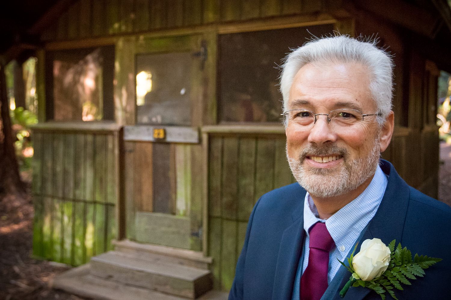 Well-dressed mature man in navy suit with boutonniere smiling in front of rustic cabin.
