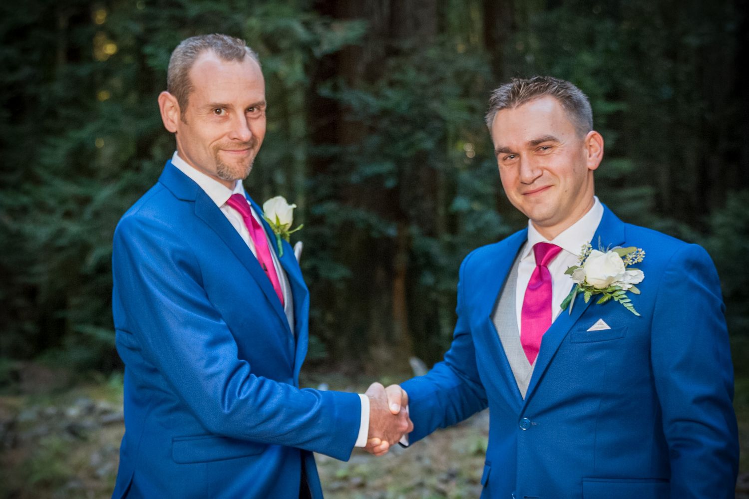 Two groomsmen in matching blue suits shaking hands during outdoor wedding ceremony.