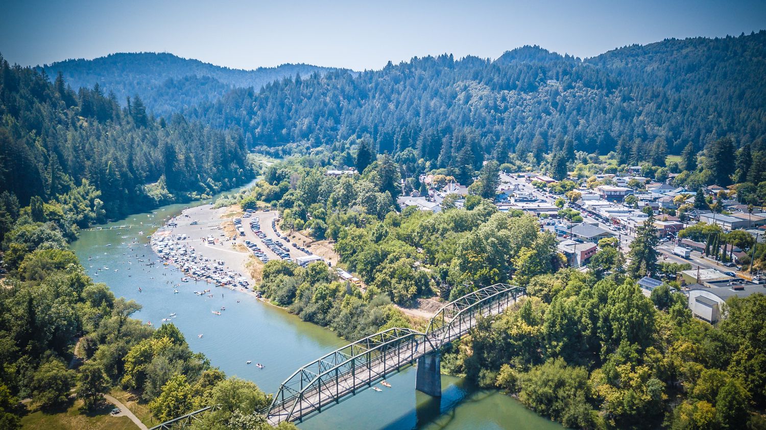 Aerial view of a small town nestled along a winding river with a footbridge connecting both sides.