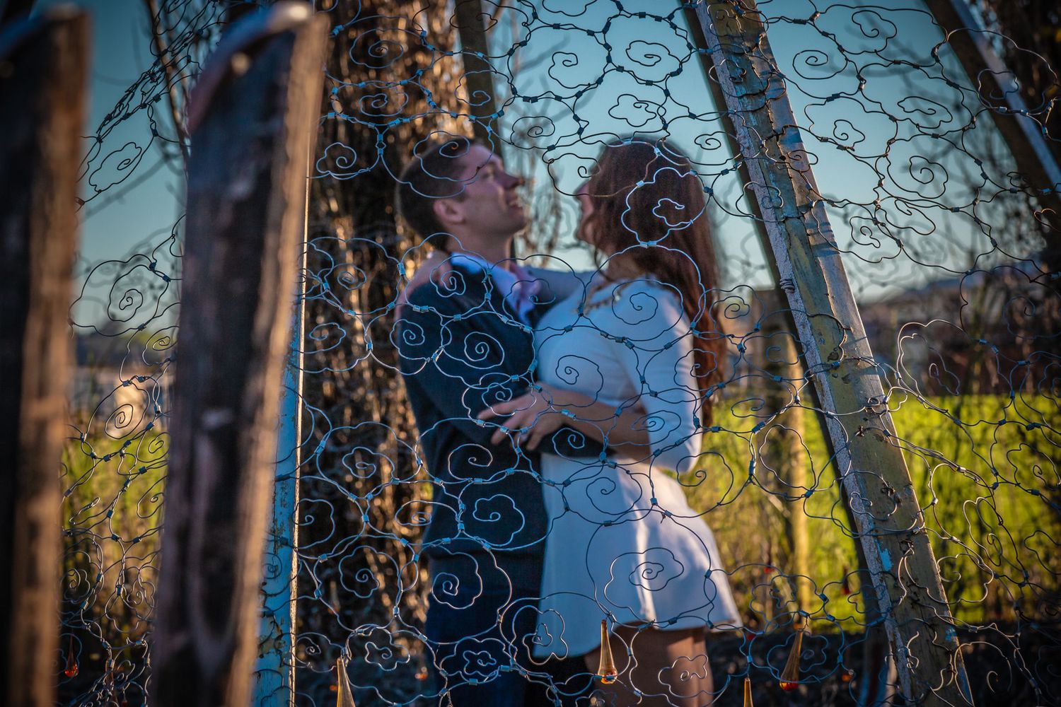 A couple shares an intimate moment through an ornate metal archway at sunset in a natural outdoor setting.