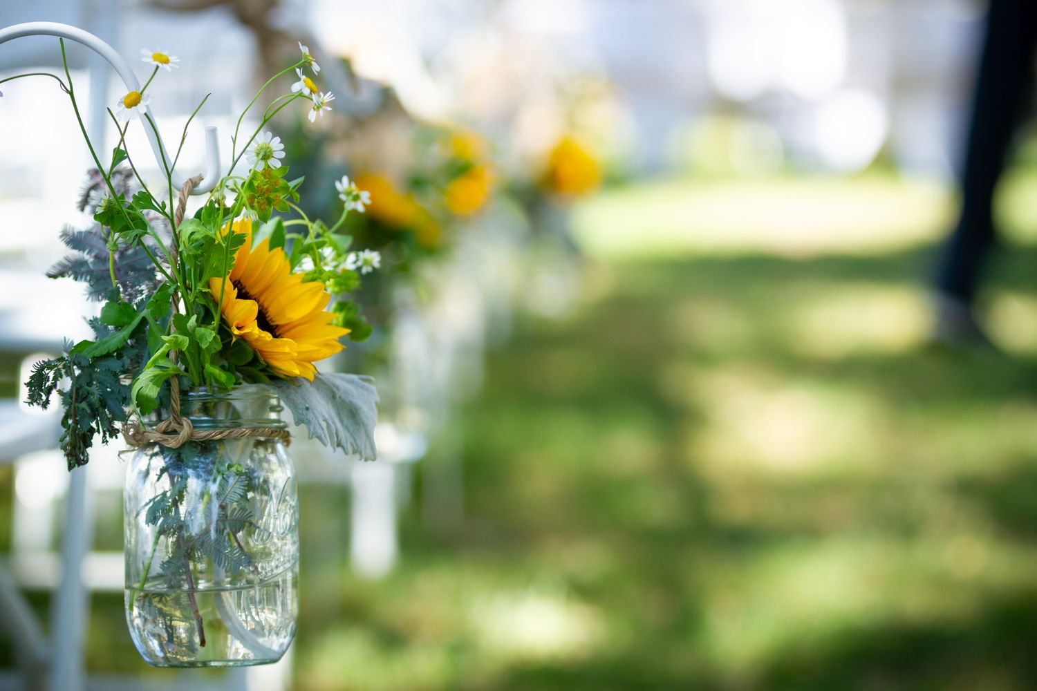 A sunflower arrangement in a hanging mason jar decorates an outdoor event with a soft blurred green background.