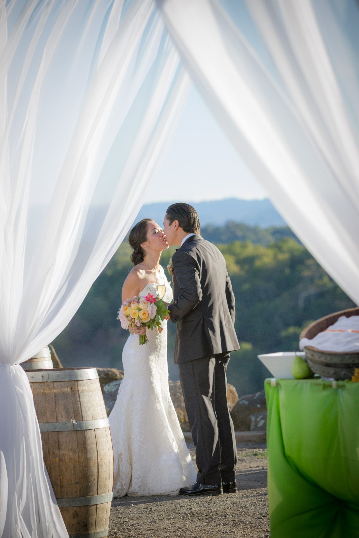 Romantic wedding couple kissing under flowing white drapery with vineyard views.
