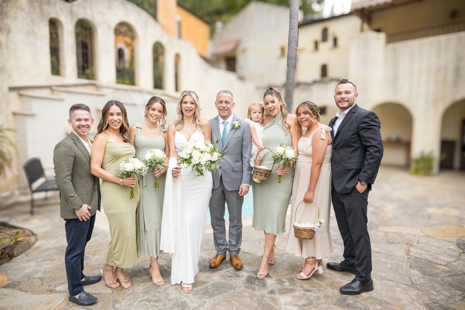 Wedding party in sage green dresses poses at Mediterranean-style venue.