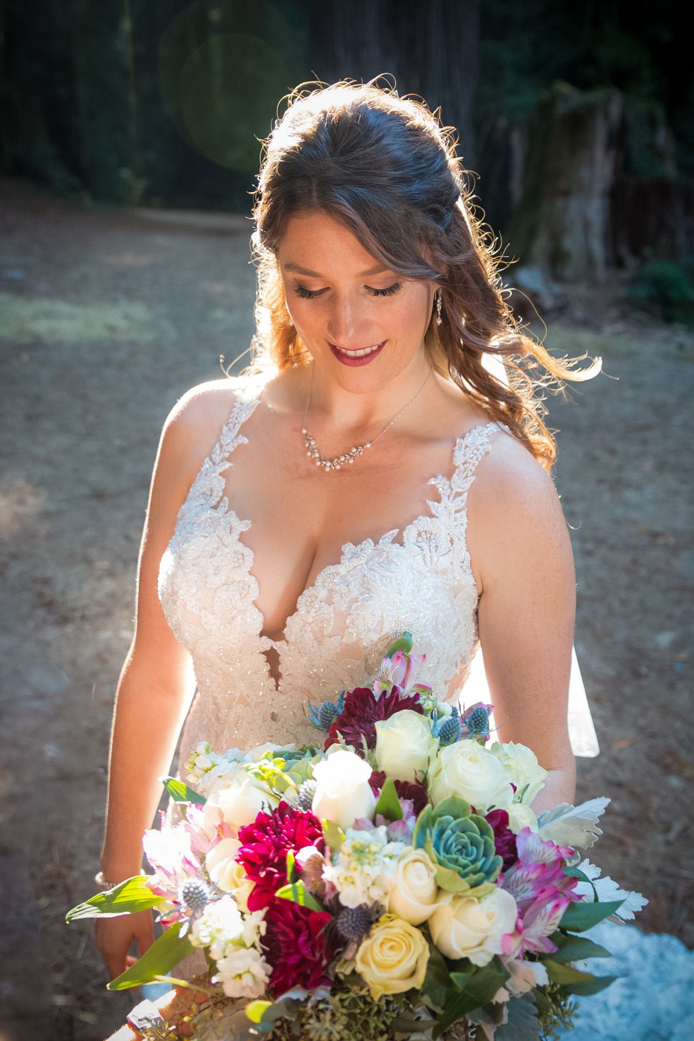 Bride in detailed lace gown holds colorful bouquet in sunlit forest setting.