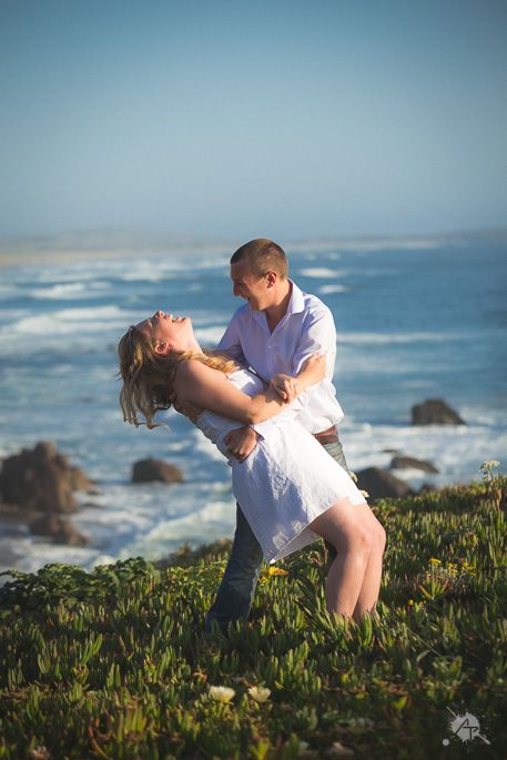 A romantic couple dips and embraces on a grassy cliff overlooking a dramatic ocean coastline at sunset.