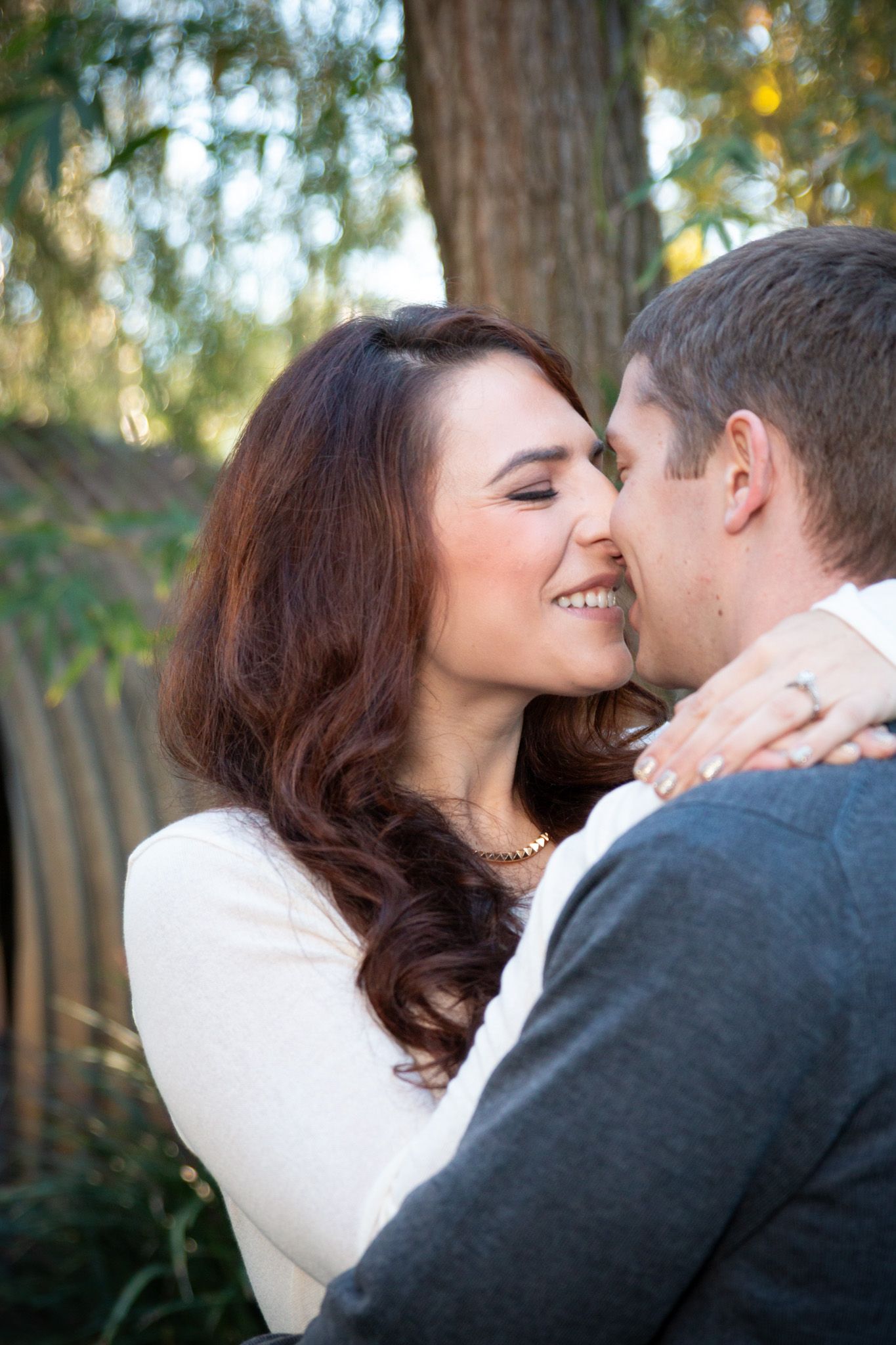 A romantic outdoor portrait of a couple embracing and smiling in a natural setting with trees in the background.