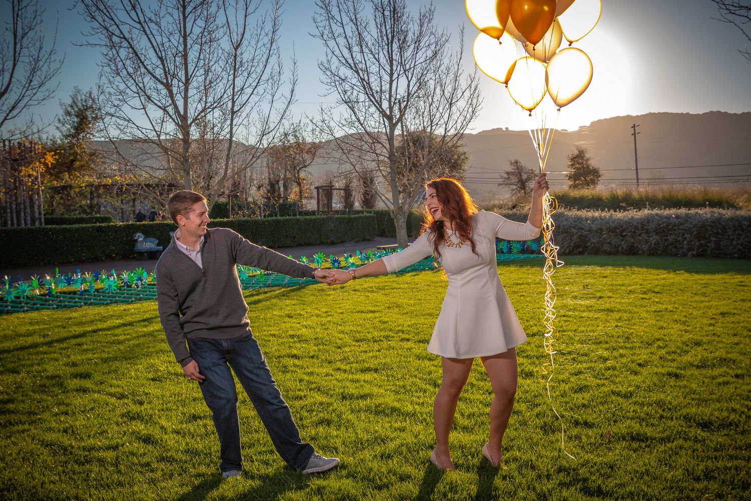 A playful couple dances together at sunset in a backyard with mountains and golden balloons in the background.