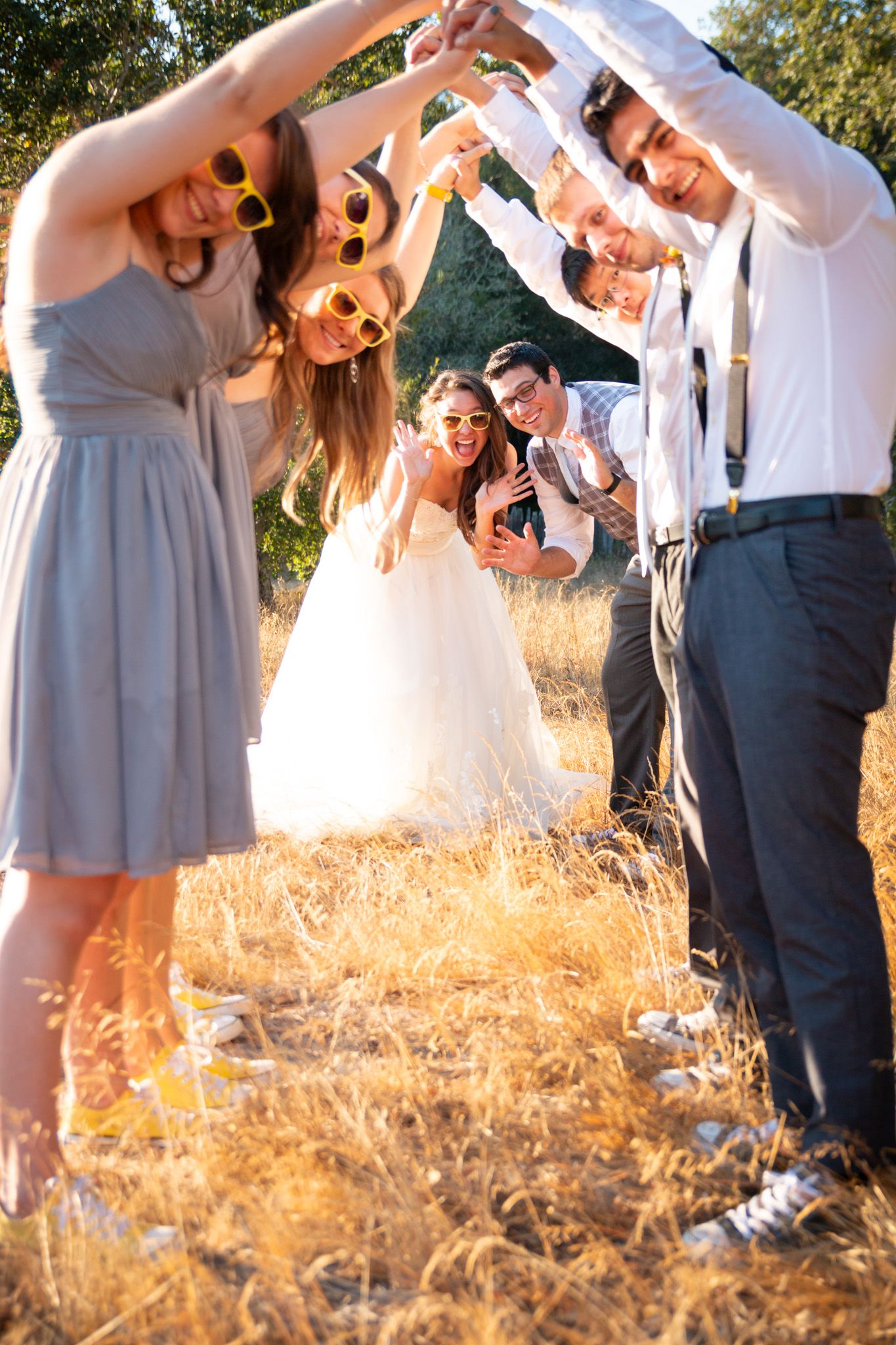 Wedding party forms archway with arms while celebrating in sunlit field.