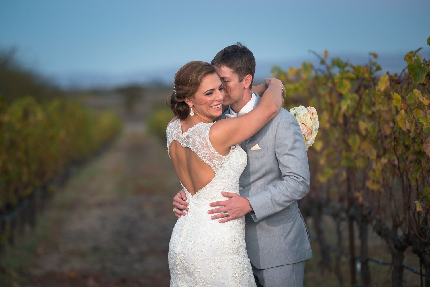 Couple embraces together in formal wedding attire among vineyard rows at dusk.