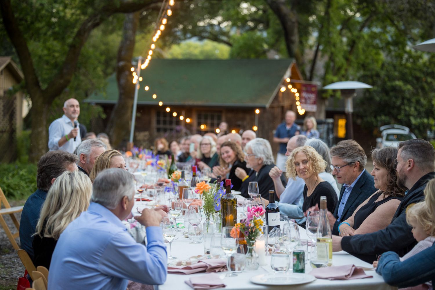 Long tables set for an elegant outdoor dinner party with string lights overhead and guests seated.