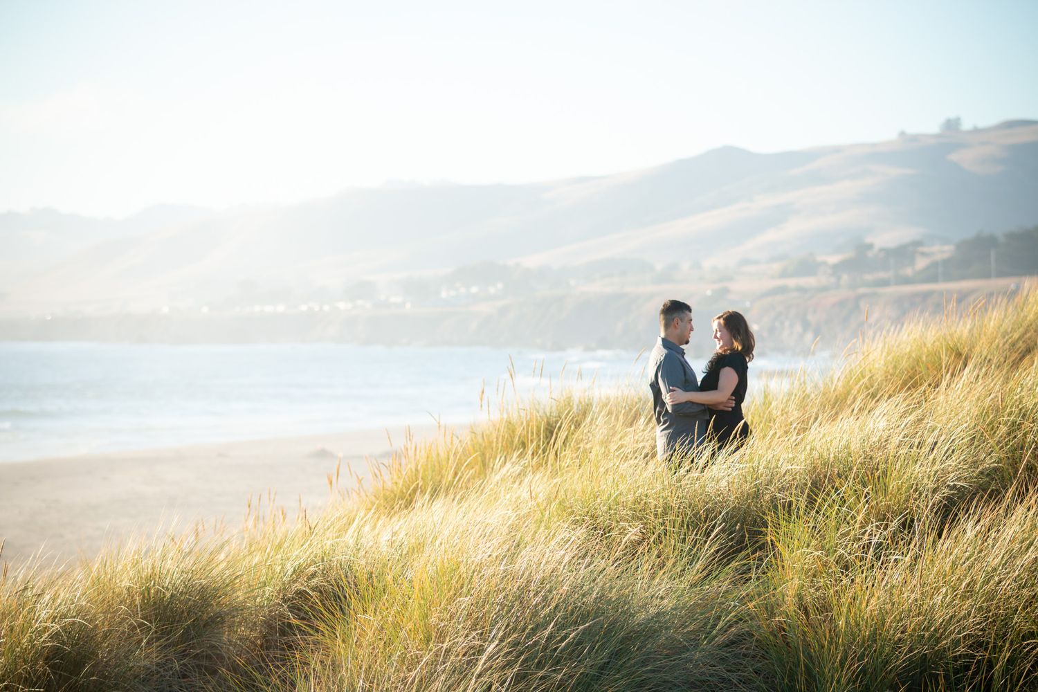 Couple embracing in tall beach grass with coastal cliffs and ocean in background.