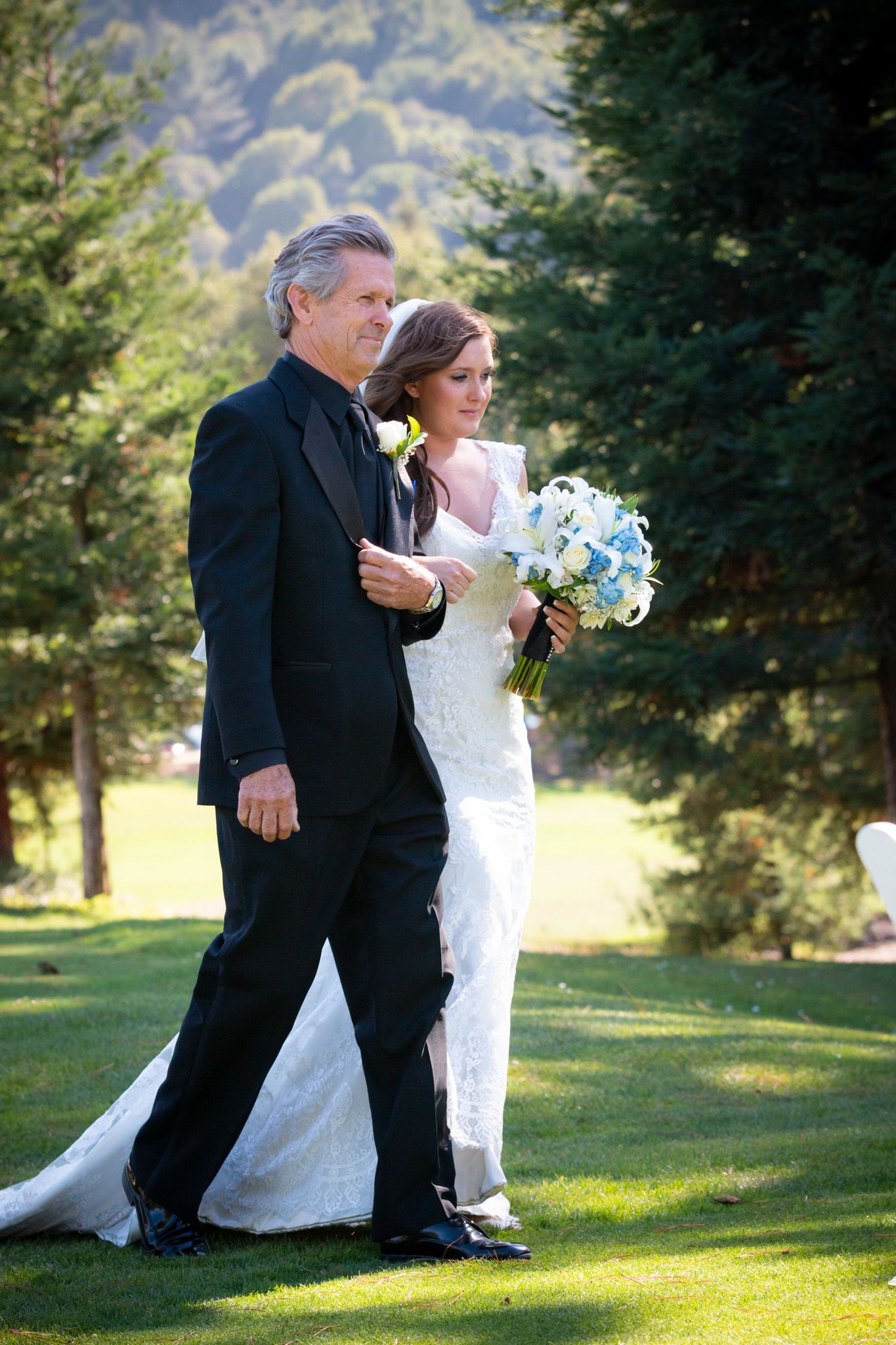 A couple walks across a sunny lawn during a wedding ceremony with mountains in the background.