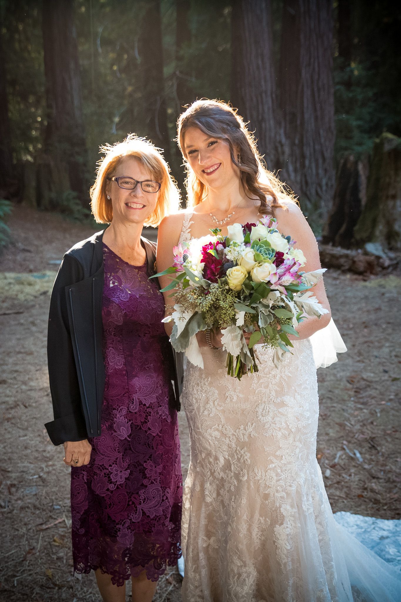 Bride in lace gown shares tender moment with wedding guest in purple dress.