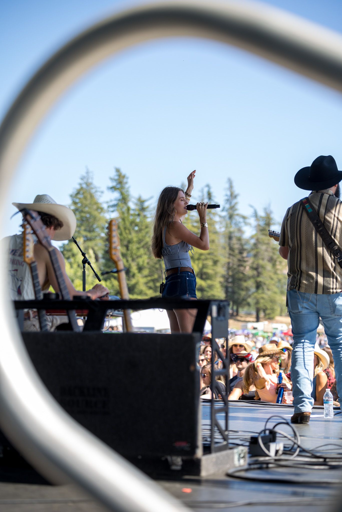 Country music performers on outdoor stage with microphones and instruments at summer festival with pine trees in background.