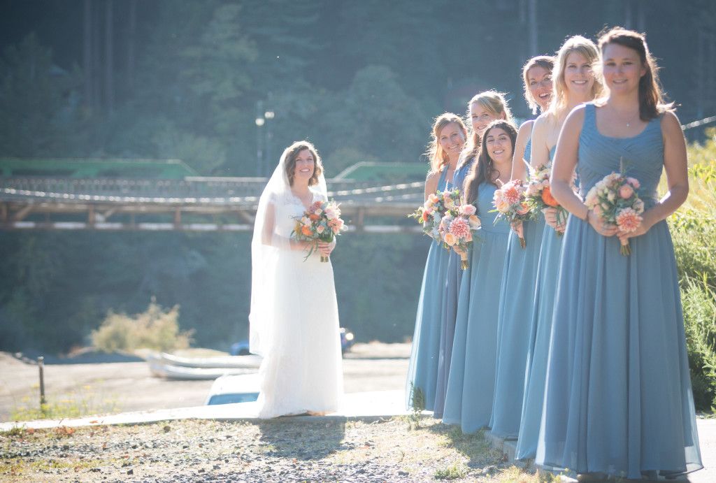 Wedding party in light blue bridesmaid dresses standing by a lake on a sunny day.