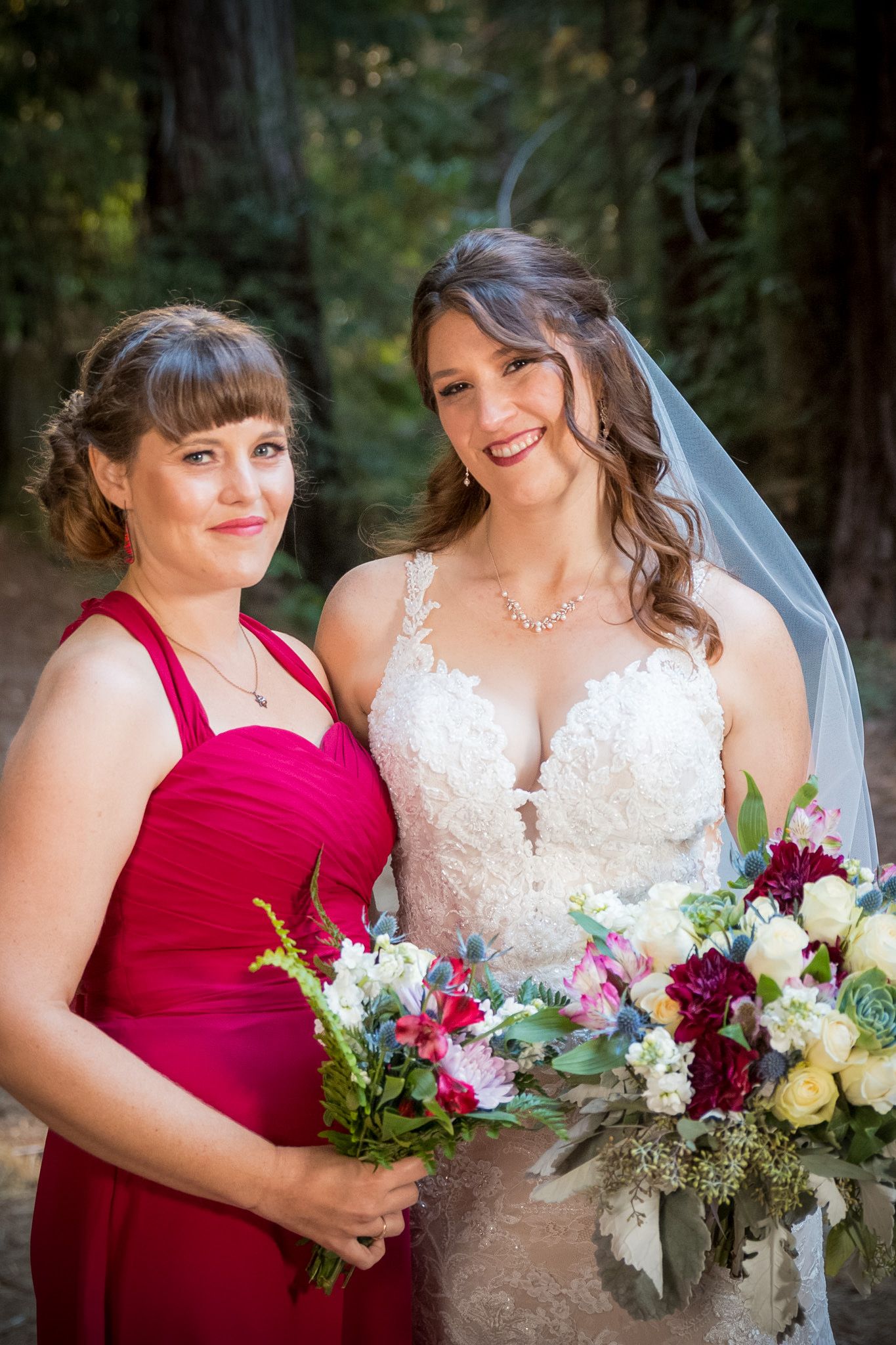 Bride in lace dress and bridesmaid in red pose with colorful bouquets in forest setting.