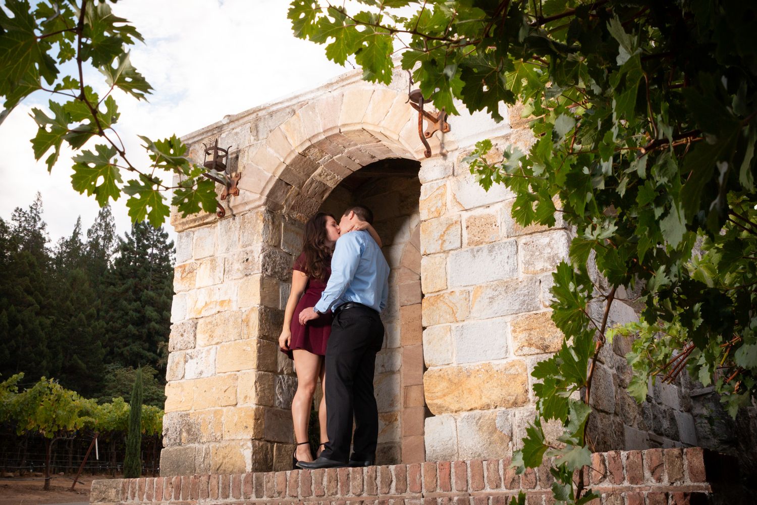 Romantic couple standing in stone archway framed by green leaves.
