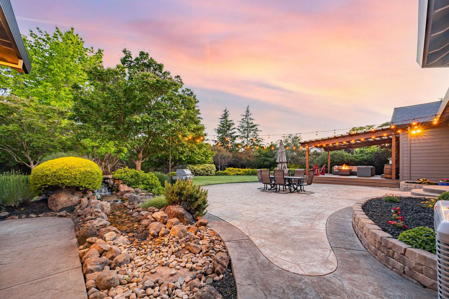 Curved stone patio with landscaped garden beds and pergola creates an inviting outdoor entertaining area at dusk.