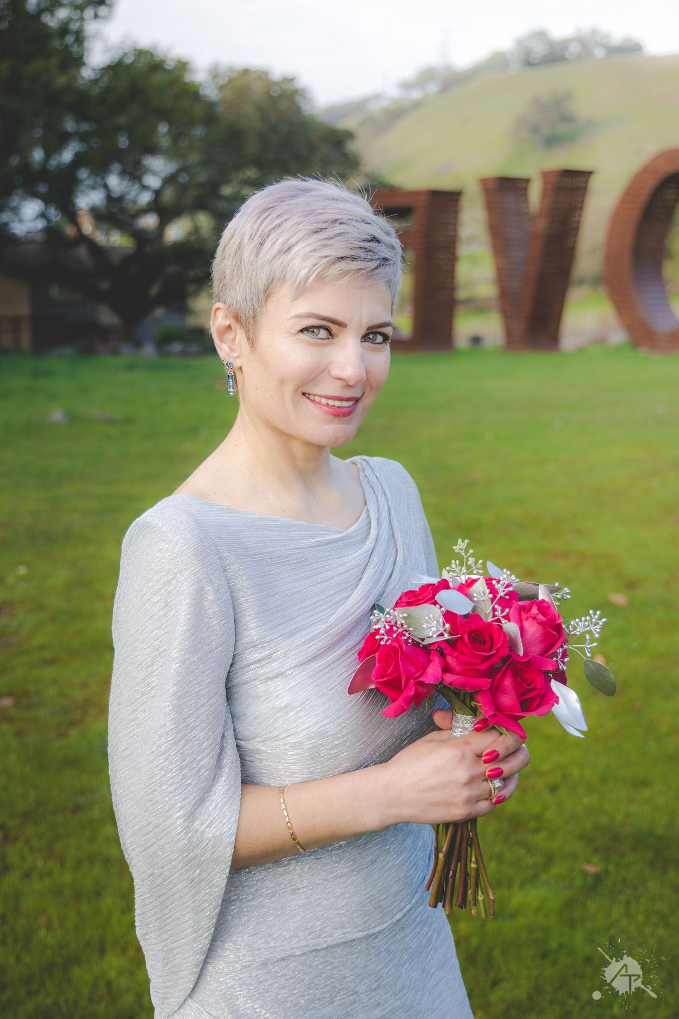 Someone in a silver grey dress holds a vibrant pink and red floral bouquet while posing outdoors near large sculptures.